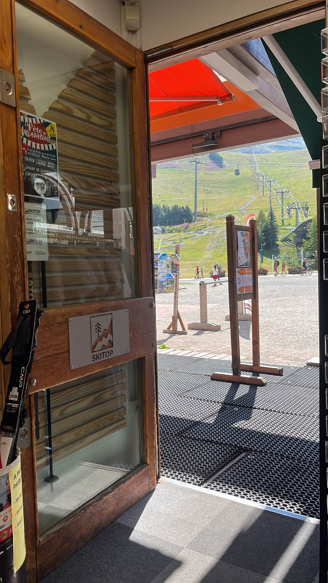 Entrée du magasin de location SKITOP ouvrant sur une station de montagne verdoyante avec ses remontées mécaniques en été.