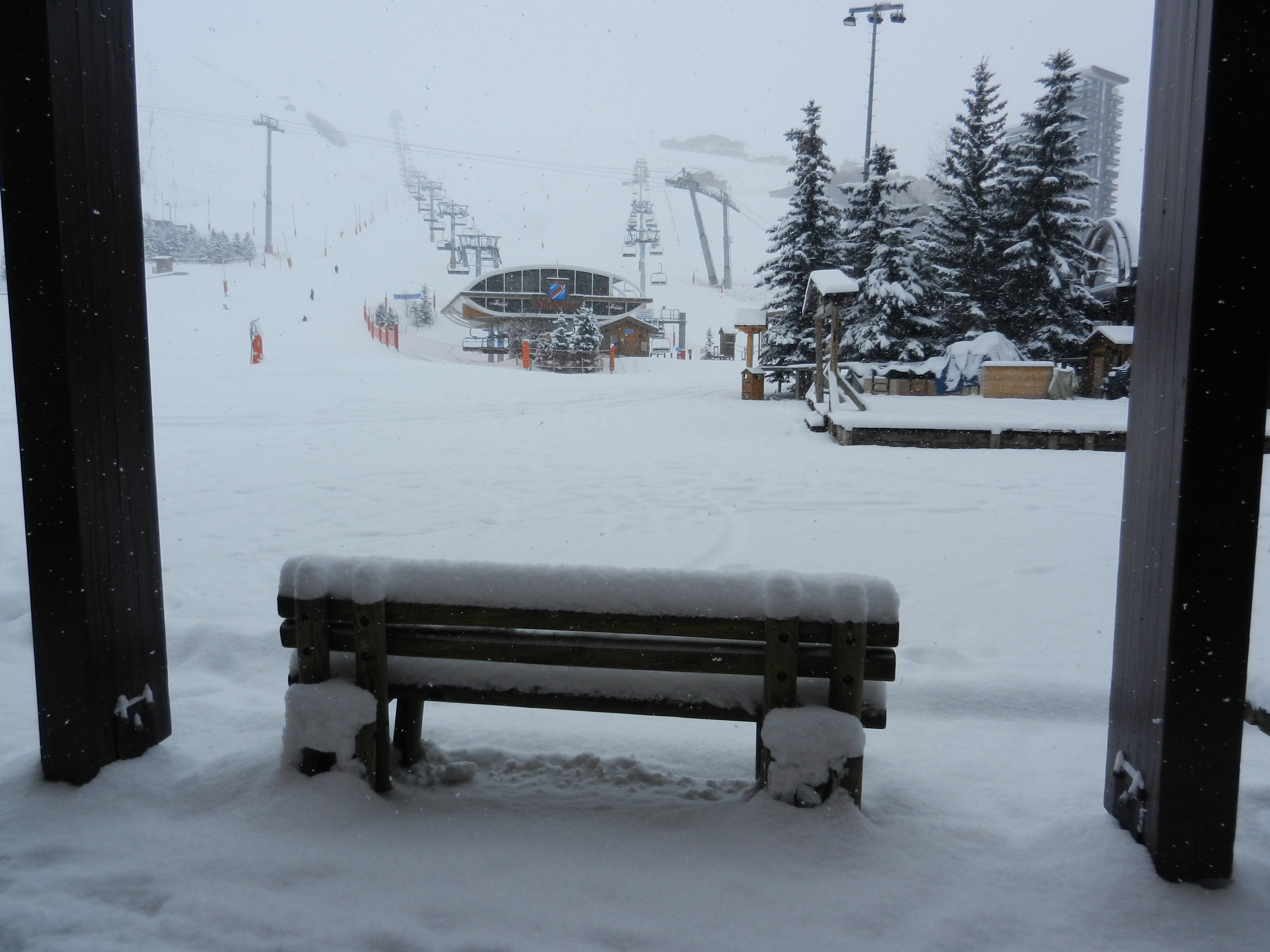 Banc en bois recouvert d'une épaisse couche de neige face aux pistes et aux remontées mécaniques d'une station de ski sous une chute de neige.